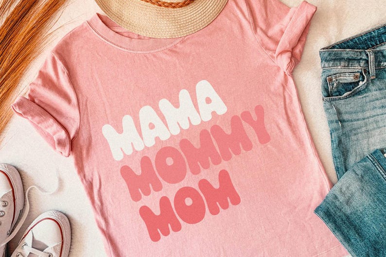 May include: A light pink t-shirt with the words "MAMA MOMMY MOM" in white and pink bubble letters. The shirt is folded, with a straw hat and blue jeans visible in the background. White sneakers are also in the image.