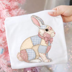 Könnte beinhalten: Ein weißes quadratisches Kissen mit einem Patchwork-Hasen-Design. Der Hase hat eine Mischung aus floralen, Vichy- und unifarbenen Flicken in Rosa, Blau und Creme. Er hat eine rosa Schleife und Ohren, ideal für Osterdekorationen.