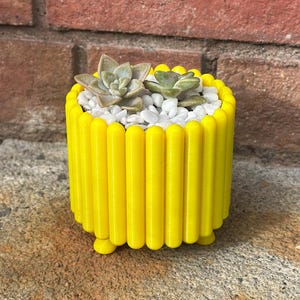 Op de afbeelding: Een heldergele cilindrische plantenbak met een geribbeld ontwerp, met kleine vetplanten en witte kiezels. De plantenbak staat op een stenen oppervlak, met een bakstenen muur op de achtergrond. De plantenbak heeft kleine voetjes.