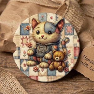 May include: A round car coaster featuring a cartoon cat wearing a hoodie and holding a teddy bear. The coaster has a patchwork design with flowers and stars. The cat's hoodie says "MADE FROM SCRAPS". A tag says "Handmade with Love".