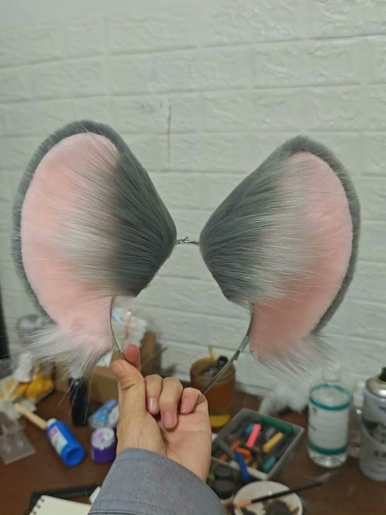 May include: A headband featuring two large, upright animal ears made of faux fur. The ears have a soft, light pink interior and a dark grey exterior with long, textured fibers. The headband frame is a thin, metallic wire connecting the two ears.