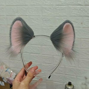 May include: A headband featuring two upright, pointed animal ears made of long, fluffy gray faux fur with soft pink inner lining. The ears are attached to a thin, flexible black plastic band, creating a distinct, textured appearance.