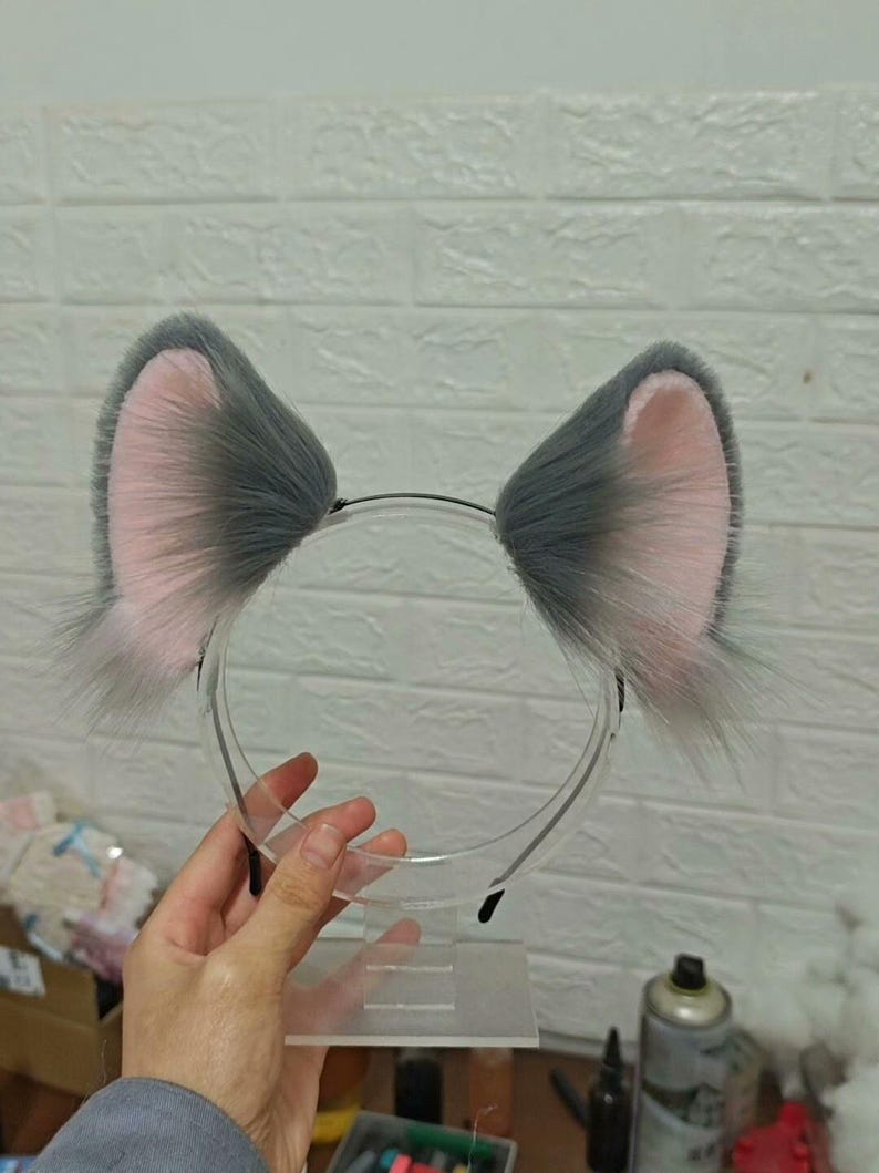 May include: A headband featuring two upright, pointed animal ears made of long, fluffy gray faux fur with soft pink inner lining. The ears are attached to a thin, flexible black plastic band, creating a distinct, textured appearance.