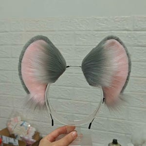 May include: A headband featuring two large, upright animal ears made of faux fur. The ears have a gradient color pattern, transitioning from a soft pink interior to a dark gray outer edge, with long, wispy gray fur fibers extending from the base.