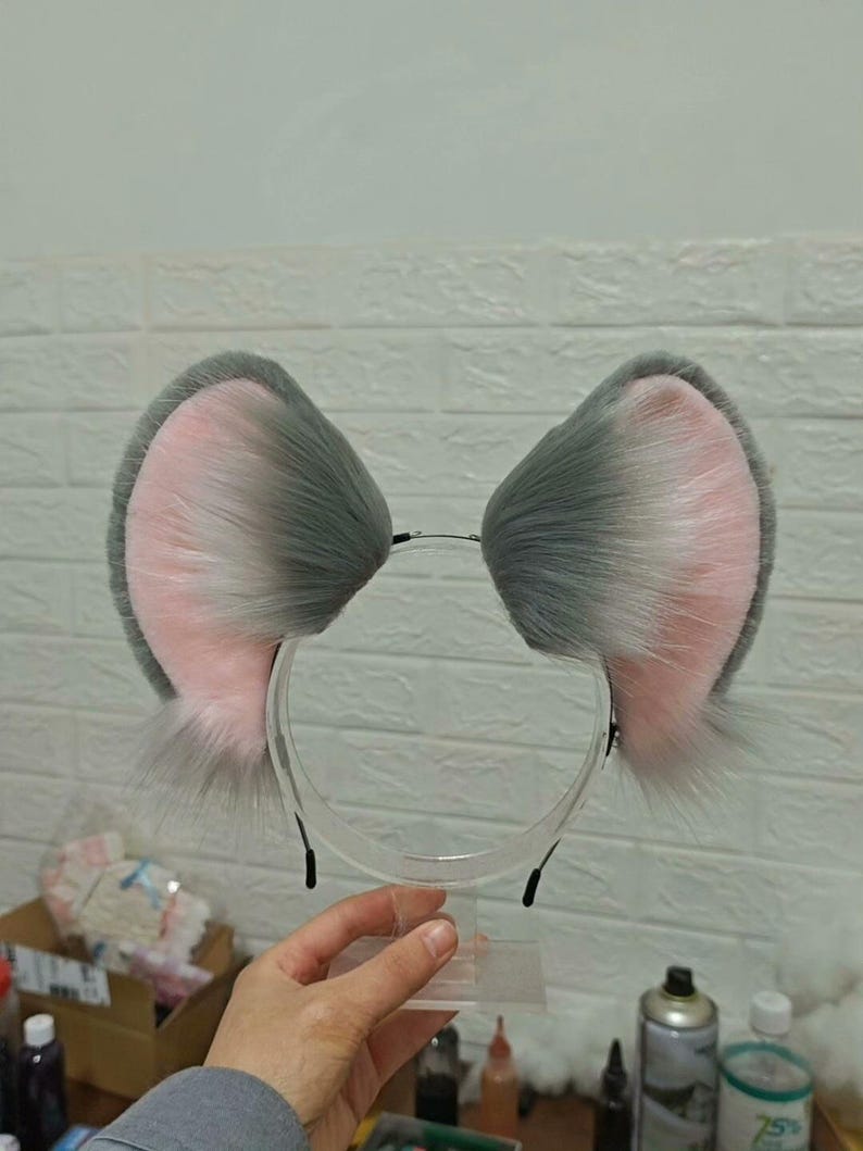 May include: A headband featuring two large, upright animal ears made of faux fur. The ears have a gradient color pattern, transitioning from a soft pink interior to a dark gray outer edge, with long, wispy gray fur fibers extending from the base.