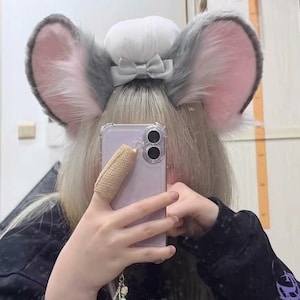 May include: A pair of plush, gray and pink animal-style ears attached to a headband, featuring a central white puff and a small gray bow. The ears have a soft, furry texture with pink inner detailing, designed to be worn on the head as an accessory.