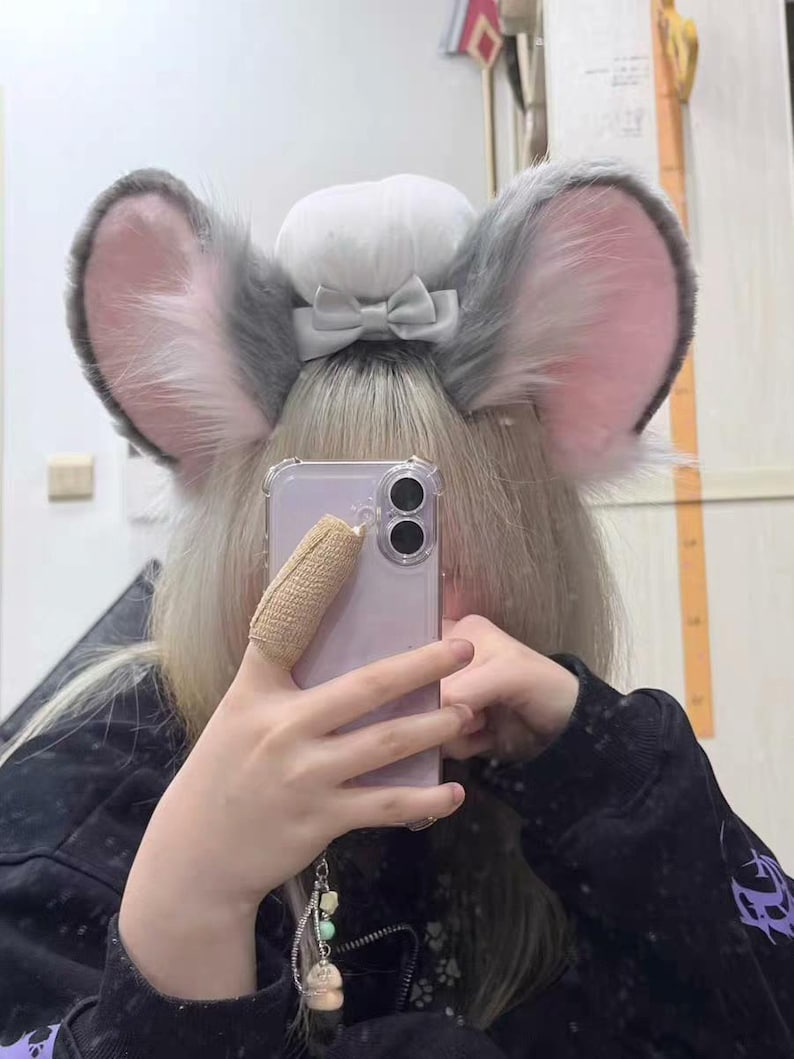May include: A pair of plush, gray and pink animal-style ears attached to a headband, featuring a central white puff and a small gray bow. The ears have a soft, furry texture with pink inner detailing, designed to be worn on the head as an accessory.