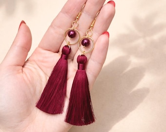 May include: A pair of dangle earrings featuring long, deep burgundy silk tassels suspended from gold-toned metal rings. Each earring includes a single matching burgundy bead centered within the circular gold frame, attached to a simple gold-toned hook wire.