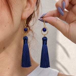Puede incluir: Un par de pendientes colgantes con un gancho de metal dorado, un marco circular dorado, una cuenta redonda de color azul intenso centrada en el círculo y una borla larga de hilo de seda azul marino que cuelga de la parte inferior.