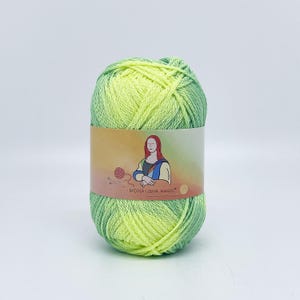 Peut inclure: Pelote de laine dégradée vert et jaune. Une étiquette entoure la pelote, avec une illustration de la Joconde et le texte "MONA LISHA MAGIC". La laine est douce et duveteuse, idéale pour les projets de tricot ou de crochet.