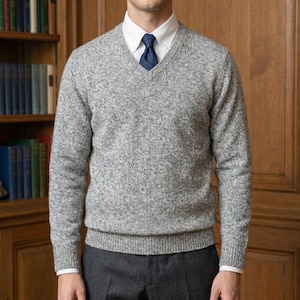 Könnte beinhalten: Ein grauer melierter V-Ausschnitt-Pullover mit langen Ärmeln, getragen über einem weißen Hemd mit Kragen und einer dunkelblauen Krawatte. Die Person trägt auch eine dunkelgraue Hose. Der Hintergrund zeigt ein Bücherregal und eine Holztür.