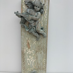 以下が含まれることがあります： リュートを演奏する彫刻されたケルビムが特徴の、長方形の壁掛け。ケルビムはブロンズ色で、色あせた装飾模様のある淡い緑色の背景に配置されています。このプラークは、風化したアンティークの外観をしています。