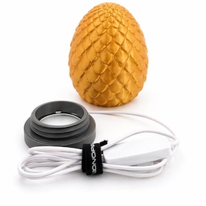 May include: A golden, egg-shaped decorative lamp with a textured, scale-like surface. Includes a gray base, a white power cord, and a black strap with the word "MONOPR" printed on it.