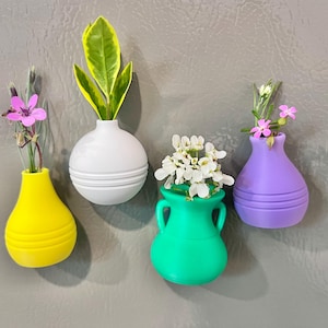 May include: Four colorful, small vases with flowers and greenery. The vases are yellow, white, green, and purple. The yellow vase has a teardrop shape. The white vase is round. The green vase has handles. The purple vase is teardrop shaped.
