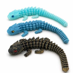 May include: Three articulated lizard figurines with segmented bodies and spiky scales. The top lizard is translucent light blue with blue eyes, the middle is solid blue with yellow eyes, and the bottom is dark bronze with red eyes.
