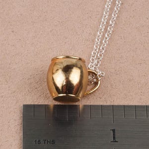 May include: A gold-toned mug pendant necklace with a silver chain. The mug is a simple design with a rounded body and a small handle.