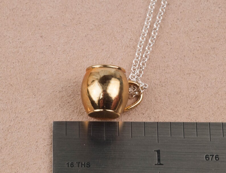 May include: A gold-toned mug pendant necklace with a silver chain. The mug is a simple design with a rounded body and a small handle.
