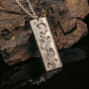 May include: A silver pendant necklace with a rectangular charm featuring an intricate design of stylized vines and leaves.