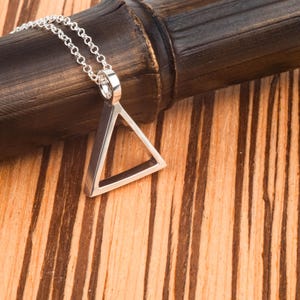 May include: A silver-toned necklace featuring a hollow triangular pendant suspended from a delicate link chain. The pendant has a smooth, polished metallic finish and a geometric shape, with the chain attached to the top corner of the triangle.