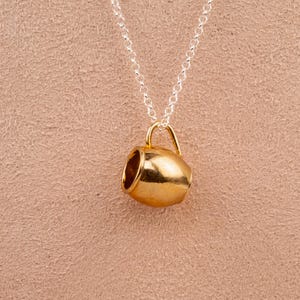 May include: A gold-toned mug pendant necklace on a silver chain. The mug is a simple design with a rounded body and a small handle.