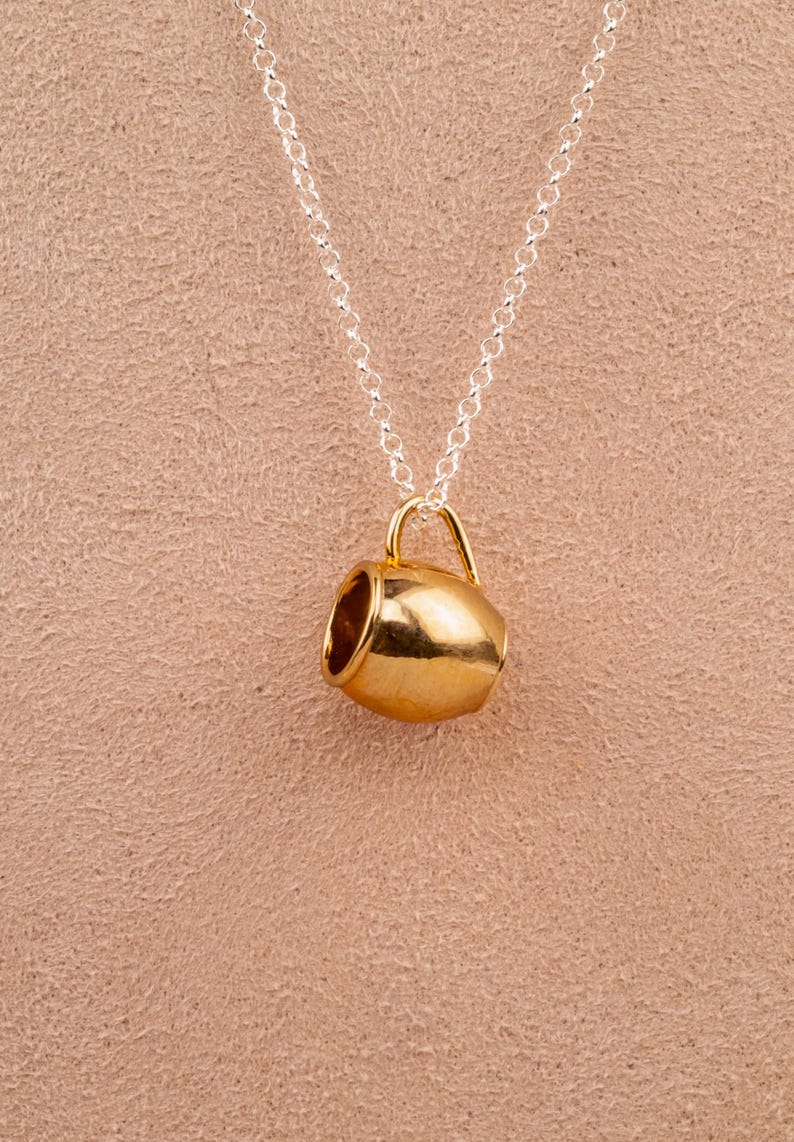 May include: A gold-toned mug pendant necklace on a silver chain. The mug is a simple design with a rounded body and a small handle.