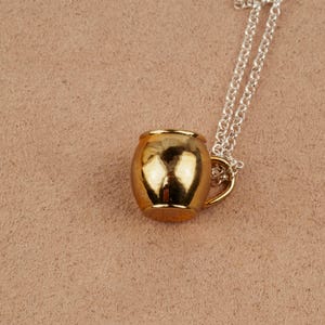 May include: A gold-toned mug pendant necklace on a silver chain. The mug is a simple design with a rounded body and a small handle.