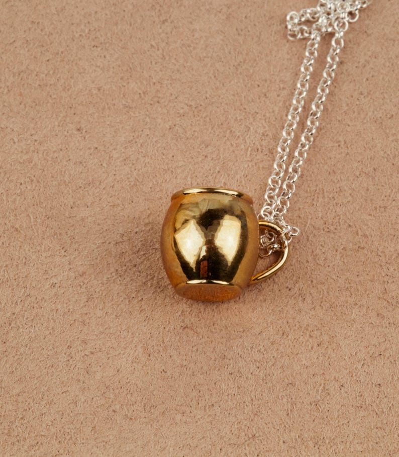 May include: A gold-toned mug pendant necklace on a silver chain. The mug is a simple design with a rounded body and a small handle.