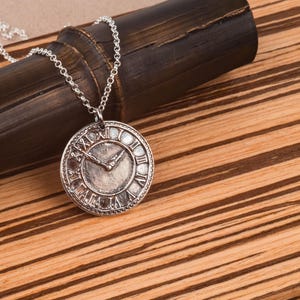 May include: A silver necklace with a round clock face pendant. The clock face has Roman numerals and a silver hand.