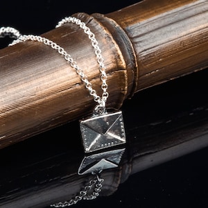 May include: A silver-colored necklace featuring a pendant shaped like an envelope. The pendant is detailed with small rivets and a textured surface. The chain is delicate and hangs against a dark background.