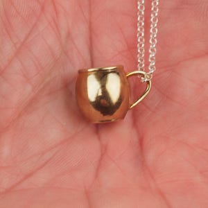 May include: A gold-colored metal mug pendant on a silver chain necklace. The mug has a rounded shape and a small handle.