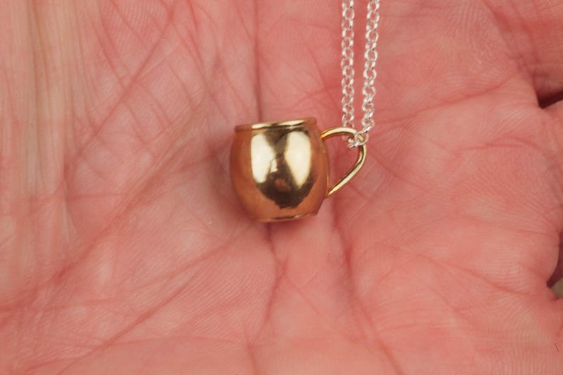 May include: A gold-colored metal mug pendant on a silver chain necklace. The mug has a rounded shape and a small handle.