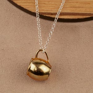 May include: A gold-colored mug pendant on a silver chain necklace. The mug is a simple design with a rounded body and a small handle.