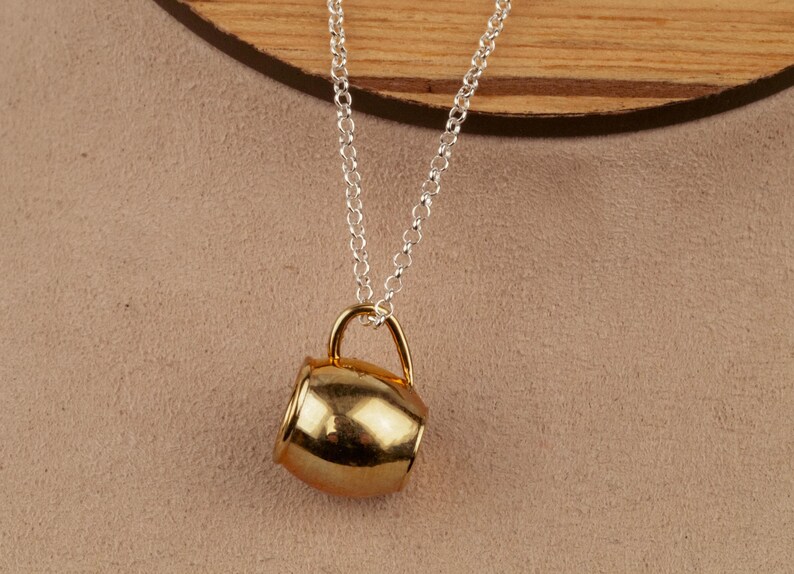 May include: A gold-colored mug pendant on a silver chain necklace. The mug is a simple design with a rounded body and a small handle.