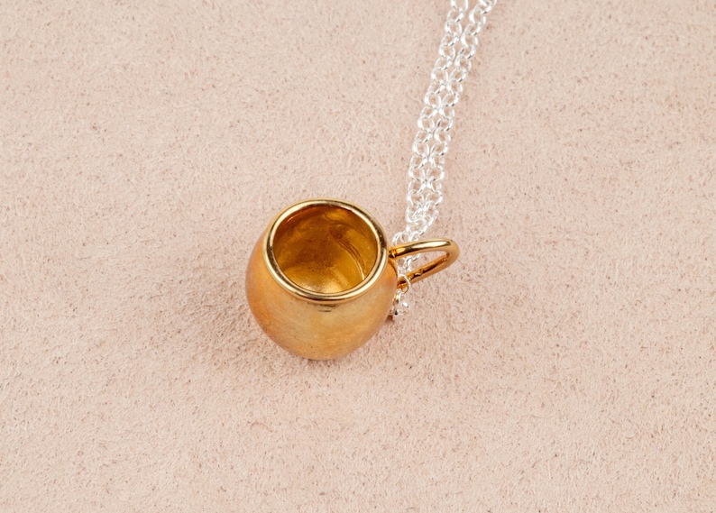 May include: A gold-colored mug pendant necklace with a silver chain. The mug is a small, detailed replica of a traditional mug.