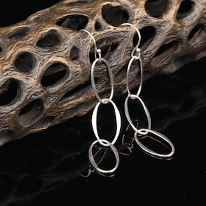 May include: Silver dangle earrings with multiple oval-shaped links. The earrings are hanging from a wooden background.