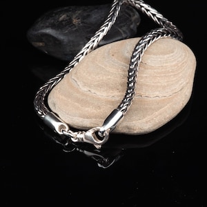 May include: A silver-colored braided chain necklace with a lobster clasp, resting on a light-colored stone. The necklace is made of interwoven links, reflecting light. A dark stone is in the background, set against a black surface.