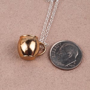 May include: A gold-toned mug pendant necklace on a silver chain. The pendant is about the size of a dime.