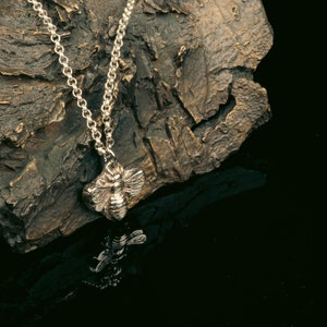 May include: A silver bee pendant necklace on a silver chain. The pendant is detailed and realistic, with wings, legs, and a stinger. The necklace is displayed on a textured, brown surface.