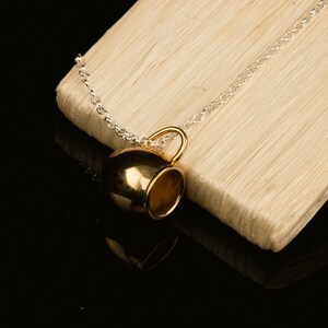 May include: A gold-colored coffee cup pendant on a silver chain necklace. The pendant is hanging from a wooden surface.