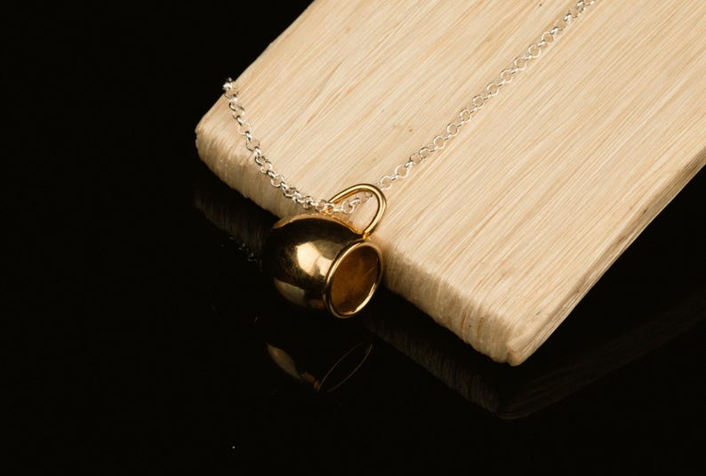 May include: A gold-colored coffee cup pendant on a silver chain necklace. The pendant is hanging from a wooden surface.