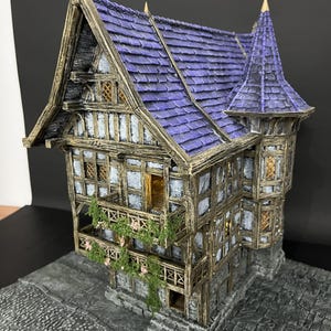 Könnte beinhalten: Ein zweistöckiges Miniatur-Fachwerkhaus mit steilem, violettem Schindeldach und spitzem Turm. Das Gebäude hat verwitterte braune Holzbalken, weiß texturierte Wände, einen Balkon mit grünen Ranken und einen Sockel in grauer Steinoptik.