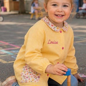 Peut inclure: Une blouse à manches longues en vichy jaune avec un col à motif floral et des poches assorties. Le vêtement présente une fermeture boutonnée rose et le prénom Louise brodé en écriture cursive rose sur la poitrine supérieure gauche.
