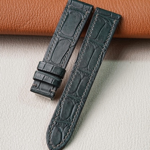 May include: A dark green watch strap featuring a textured alligator-style embossed pattern. The strap consists of two pieces, one with two sliding loops and both finished with light-colored stitching along the edges, showcasing a matte finish.