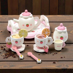 Pode incluir: Um conjunto de chá de madeira em branco e rosa. O conjunto inclui uma chaleira, xícaras, pires, colheres e uma açucareira. A chaleira e as xícaras são decoradas com desenhos de morangos e flores. O conjunto inclui uma bandeja e fatias de limão e laranja.