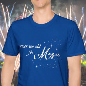 Könnte beinhalten: Königsblaues T-Shirt mit dem weißen Schriftzug "never too old for Magic" und Sternen. Das T-Shirt hat einen Rundhalsausschnitt und scheint aus einem weichen Material zu sein. Der Hintergrund zeigt ein verschwommenes Bild von Feuerwerk.