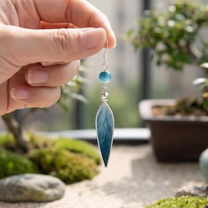 Op de afbeelding: Een zilveren oorbel met een blauwe, veervormige hanger en een kleine blauwe kraal. De oorbel wordt getoond tegen een achtergrond van bonsai-bomen en mos, wat een natuurlijke esthetiek creëert. Het veermotief heeft blauwe tinten.