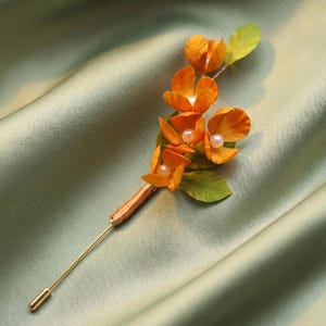 May include: A floral brooch with orange blossoms, pearl accents, and green leaves. The brooch has a gold-coloured pin, and the flowers are arranged on a brown stem. The background is a light green, silky fabric.
