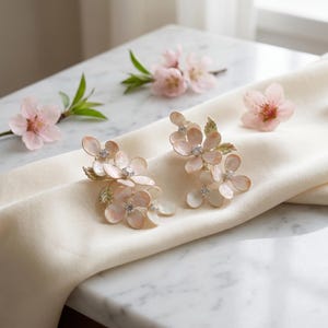 May include: A pair of floral earrings featuring pale pink, iridescent petals with small green leaves and clear crystal centers. The earrings are arranged on a soft, cream-colored fabric, accompanied by three small, light pink blossoms.