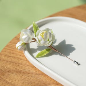 May include: A white floral lapel pin with three blossoms and green leaves. The pin is set on a white ceramic plate and has a silver-coloured pin backing. The flowers have yellow centres and are arranged on a brown stem.