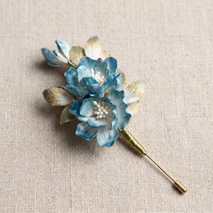 Può includere: Una spilla da bavero floreale artigianale con fiori blu e bianchi, centri bianchi e dettagli dorati. La spilla ha un lungo e sottile spillo color oro per il fissaggio. Le foglie sono un mix di verde e oro.